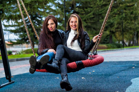 Two beautiful young brunette and brown-haired women with long hair sit on a round swing in the park...