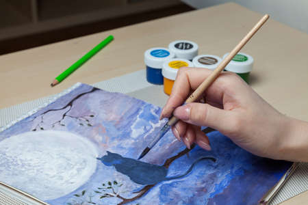 Female hands hold a brush and a cover from a watercolor jar and mix paints against the background of...