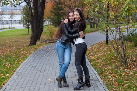 Two young women - girlfriends, students in fall on the embankment stand on the promenade against the background of trees with yellow-red leaves and pose hugging in leather jackets, sweaters and jeans.の写真素材
