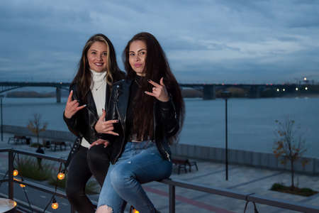 Beautiful brunette young girlfriends pose for photo in evening at embankment, sitting on railing of summer roof of a cafe-veranda with garlands and light bulbs against background of river and bridge.の写真素材