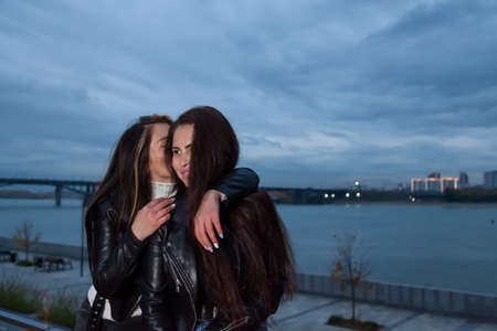 Young brunette friends with long hair hug in the evening at sunset on the embankment against the backdrop of the night city and the bridge of the opposite bank. Female friendship or a couple in love.の写真素材