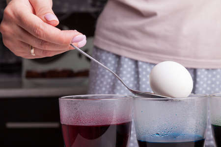A white boiled egg on an iron spoon in women's hands before dipping into a transparent glass with...
