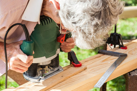 An elderly man at his summer cottage in a panama hat works with a green manual milling machine over lumber against a background of bushes.の写真素材