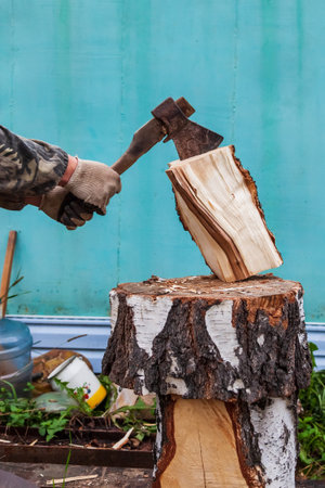 A log split in half with an ax on a deck with male hands in cloth gloves and a camouflage jacket in the backyard. Household work to prepare for the winter.の写真素材