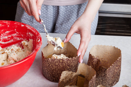 Close-up of brown paper baking tins filled with raw raisin dough and leveling it with a cooking muffin spoon at home.の写真素材