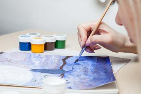 A close-up of female hands over a painted picture holding a brush and a cover for watercolors on which the colors are mixed next to the eraser and a simple pencil. A hobby of art.の写真素材