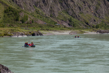 A team of people rafting in equipment, lifejackets and yellow helmets on a blue inflatable boat along a mountain river with rapids with raised oars.のeditorial素材