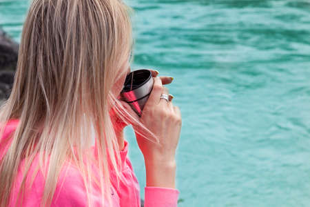 a young beautiful blonde girl in a pink suit with shorts sits on a large stone near the river in the altai mountains and drinks hot tea from an iron mug while relaxingの写真素材