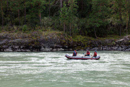 Novosibirsk, Russia - 08.14.2020: A team of people rafting in orange life jackets on rubber boat of blue and yellow colors along a mountain river against the background of a rocky shore with a forestのeditorial素材