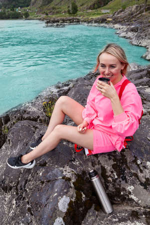 a young beautiful blonde girl in a pink suit with shorts sits on a large stone near the river in the altai mountains and drinks hot tea from an iron mug while relaxingの写真素材
