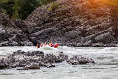A team of people rafting in orange life jackets on an rubber boat of blue and yellow colors along a...