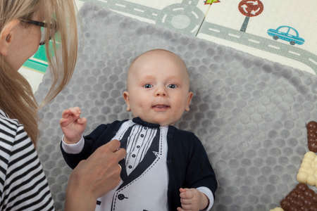 Young mom dresses her baby little boy in gentleman's outfit, light interior, concept of motherhood