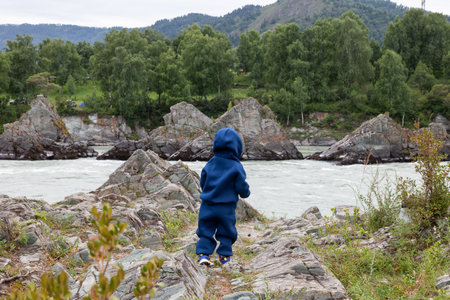 Little baby in blue sport suit playing on the bank of the river katun with Altai mountain peaks in the background. vacation for family with childの写真素材