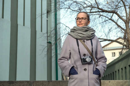 Blonde woman with glasses and with a camera standing in a coat on the street