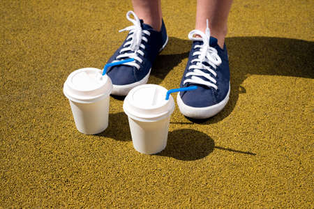 Female feet in blue sneakers and two cups of coffee on yellow coating on a sunny dayの写真素材