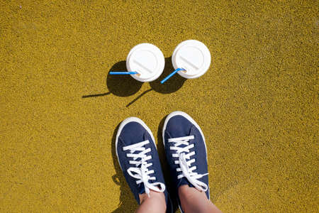 Female feet in blue sneakers and two cups of coffee on yellow coating on a sunny dayの写真素材