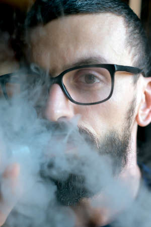 Bearded man with glasses smoking vaporizer and blows smokeの写真素材