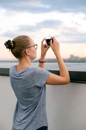 Girl blonde with glasses photographed on a white mobile phone on the roof of the house. Summer daytimeの写真素材