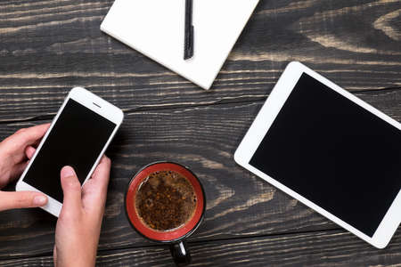 Hand with white phone, black and red cup of coffee, white tablet, white notepad with black pen on the aged wooden dark backgroundの写真素材