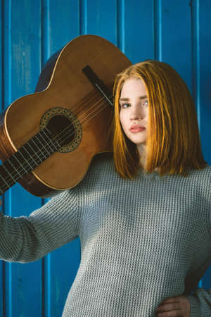 Red-haired girl standing with acoustic guitar on his shoulder against the backdrop of a blue wall. Redhead.の写真素材