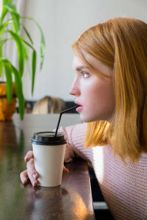 Red-haired girl sitting at a table with a cup of coffee. Redhead paper cup of coffee.