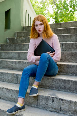 Red-haired girl sitting on the stairs with a black folder in hands. Redhead.の写真素材