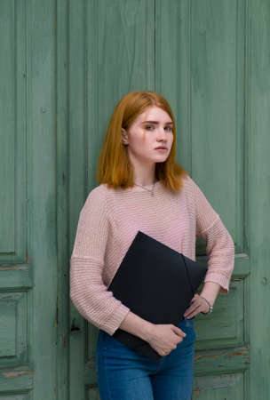 Red-haired girl standing with a black folder in hands. Red-haired girl standing with a black folder in hands on a background of green wooden wall. In the daytime.の写真素材