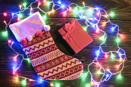 Christmas red sock and lights background. Holiday shiny garland border top view on brown wooden planks surface. Xmas tree decorations, winter holidays illumination. New Year. Red gift boxの写真素材