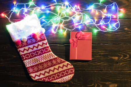 Christmas red sock and lights background. Holiday shiny garland border top view on brown wooden planks surface. Xmas tree decorations, winter holidays illumination. New Year. Red gift boxの写真素材