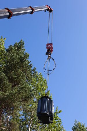 Crane hook lifts up the power transformer against the green forest