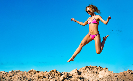 Photo of beautiful young model jumping, in the air, on pile of sand and blue sky backgroundの写真素材