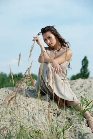 fashionable girl in a long light dress sits on a warm sandy beach and looks at the camera. Charming summer style girl, windy hair, delicate skin. Summer day and happy holidaysの写真素材