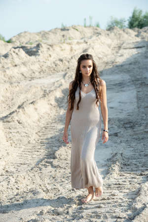 Fashionable young woman in a long dress walks on the sandの写真素材