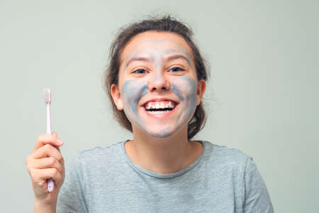 Happy teenage girl with a cosmetic mask on her face brushes her teeth and smiles cheerfully. Scrub face mask. The concept of health and beauty.の写真素材