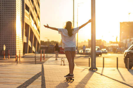 Girl on a skateboard illuminated by the rays of the setting sun. rides on a skateboard with his arms raised high, his back to the camera. Selective focus. Sunset in the cityの写真素材