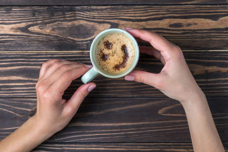 Female hands holding a cup of coffee with foam over a wooden table, top view. Latte or cappuccino...