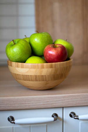 A wooden bowl with green apples and one red Apple in close-up on the kitchen table. There is a...