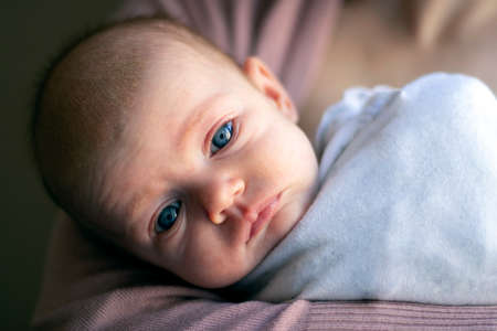 Close-up of a newborn baby with no hair and blue eyes lying on the mother's hand and looking straight. The child is wrapped in white clothes. U-turn of the head-full face.の写真素材