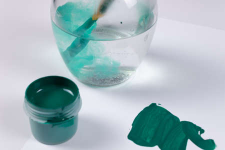 the brush is dipped into a transparent jar of water, the water is slightly colored green, next to a thick smear of green paint and an open jar of green gouacheの写真素材