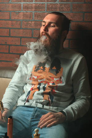 Vape. Young brutal white guy with large beard is admiting puffs of steam from the electronic cigarette.の写真素材