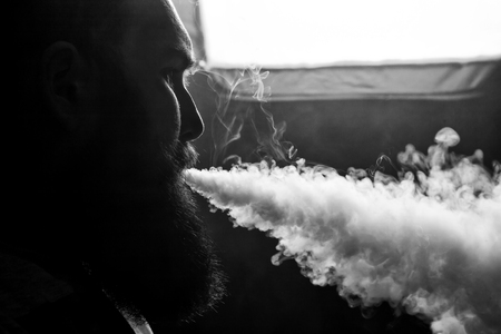 Vape. Young handsome white guy with large beard is admiting puffs of steam from the electronic...