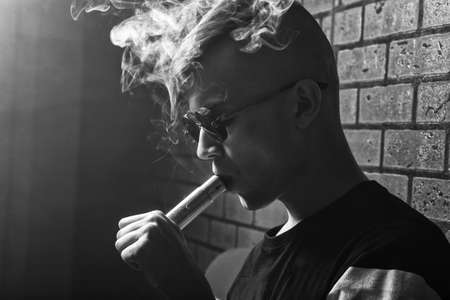 Vape. Young handsome white guy in sunglasses is admiting puffs of steam from the electronic cigarette. Vaping. Teenager listening to music. Black and white photo.の写真素材