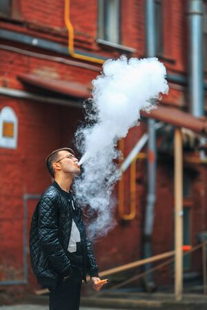 Vape. A handsome young white guy in glasses blows steam from an electronic cigarette in a vintage...