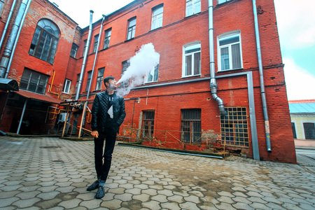 Vape. A handsome young white guy in glasses blows steam from an electronic cigarette in a vintage old red yard. Vaping.の写真素材