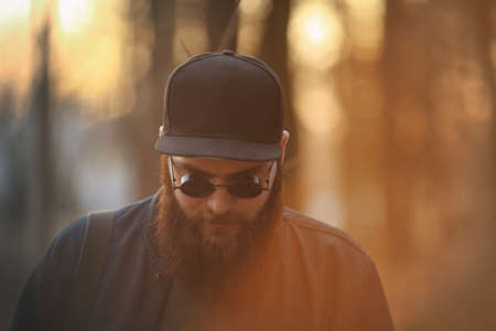 A brutal young man with a huge beard in sunglasses and a cap in the woods at sunset. Lifestyle.の写真素材