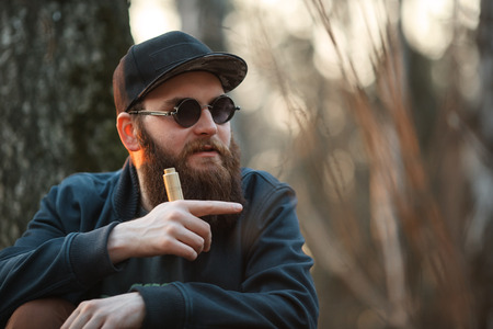 Vape. A brutal young man with a huge beard in sunglasses and a cap and with electronic cigarette in the woods at sunset. Lifestyle.の写真素材