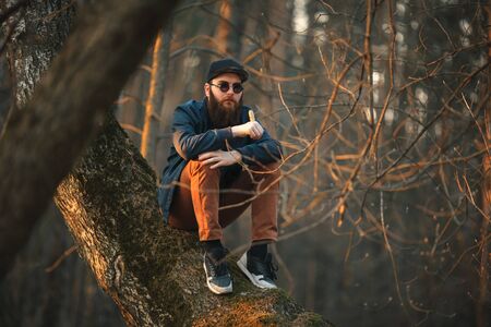 Vape. A brutal young man with a huge beard in sunglasses and a cap and with electronic cigarette in the woods at sunset. Lifestyle.の写真素材