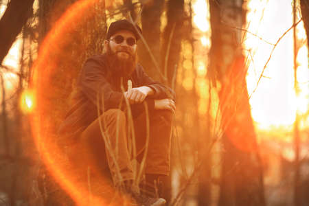 Vape. A brutal young man with a huge beard in sunglasses and a cap and with electronic cigarette in the woods at sunset. Lifestyle.の写真素材