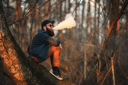 Vape. A brutal young man with a huge beard in sunglasses and a cap is smoking an electronic cigarette in the woods at sunset. Lifestyle.の写真素材