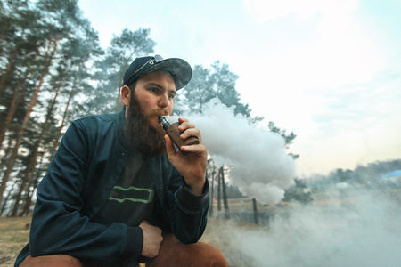 Vape. Young man with large beard in a cap smokes an electronic cigarette in the forest.